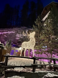 A glowing reindeer made of string lights stands amidst a snowy scene, surrounded by pine trees. The background features a rocky surface and a building, while purple lighting casts a hue over some areas. The night sky provides a dark contrast to the illuminated foreground.