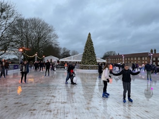 An outdoor ice rink bustling with people skating. A large Christmas tree adorned with lights stands prominently at the center. The scene is lively, with people of various ages gliding on the ice. The overcast sky looms above, while festive lights and decorations add a warm glow to the surroundings.
