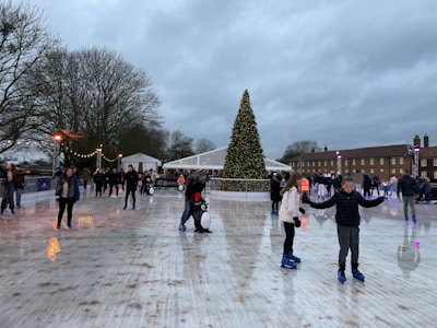 An outdoor ice rink bustling with people skating. A large Christmas tree adorned with lights stands prominently at the center. The scene is lively, with people of various ages gliding on the ice. The overcast sky looms above, while festive lights and decorations add a warm glow to the surroundings.