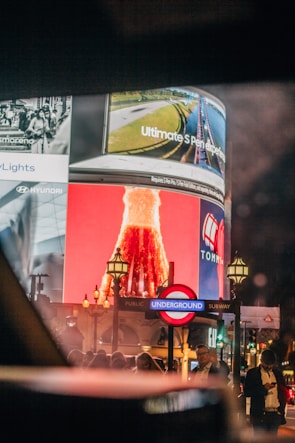 Large digital advertisements displayed on a curved screen above a busy street corner, featuring an illuminated sign for the London Underground. The scene is bustling with people and streetlights, suggesting a lively urban setting.