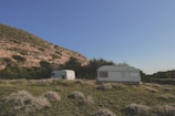 Two caravans are situated on a grassy field with sparse vegetation. They are surrounded by lush green bushes and trees. A large hill with rocky outcrops and patches of green is visible in the background under a clear blue sky.