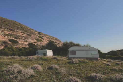Two caravans are situated on a grassy field with sparse vegetation. They are surrounded by lush green bushes and trees. A large hill with rocky outcrops and patches of green is visible in the background under a clear blue sky.
