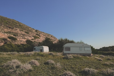 Two caravans are situated on a grassy field with sparse vegetation. They are surrounded by lush green bushes and trees. A large hill with rocky outcrops and patches of green is visible in the background under a clear blue sky.