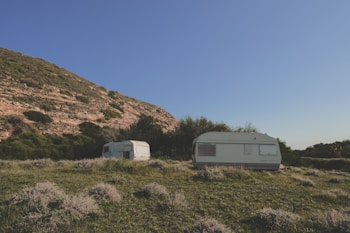 Two caravans are situated on a grassy field with sparse vegetation. They are surrounded by lush green bushes and trees. A large hill with rocky outcrops and patches of green is visible in the background under a clear blue sky.