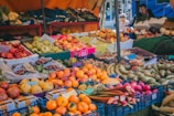 A vibrant market stall filled with a variety of fresh fruits and vegetables. Numerous crates and boxes display apples, bananas, oranges, pears, and other produce. People are seen in the background, likely shopping or conversing.