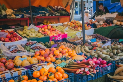 A vibrant market stall filled with a variety of fresh fruits and vegetables. Numerous crates and boxes display apples, bananas, oranges, pears, and other produce. People are seen in the background, likely shopping or conversing.