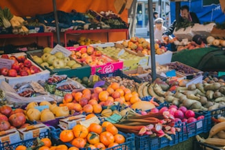 A vibrant market stall filled with a variety of fresh fruits and vegetables. Numerous crates and boxes display apples, bananas, oranges, pears, and other produce. People are seen in the background, likely shopping or conversing.