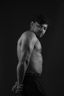 A muscular man is standing sideways against a dark background with his hands clasped behind his back. His eyes are closed, and he appears contemplative. The light emphasizes his toned physique.