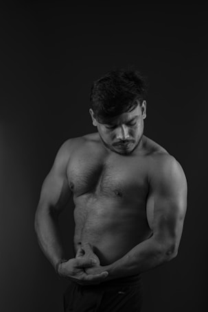 A black and white photo of a muscular male posing with a focus on his biceps and chest. His head is slightly bowed, and his expression is contemplative. The image is low-key with a dark background, highlighting the contours and definition of his muscles.