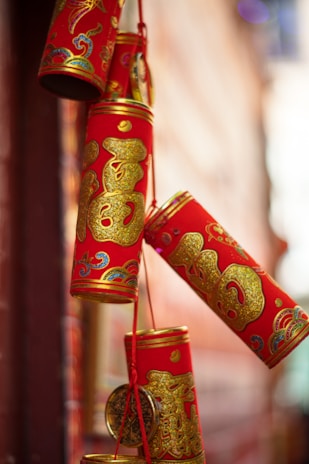 A cluster of decorative red cylinders adorned with intricate gold patterns and Chinese characters, likely representing good fortune or celebration. The cylinders are hanging by red strings, creating a festive atmosphere.