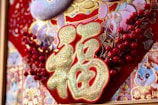 An ornate decoration with prominent gold Chinese characters, surrounded by intricate patterns featuring colorful flowers and cartoon-like animal images. Red berries and gold accents enhance the design against a predominantly red background.