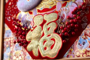 An ornate decoration with prominent gold Chinese characters, surrounded by intricate patterns featuring colorful flowers and cartoon-like animal images. Red berries and gold accents enhance the design against a predominantly red background.