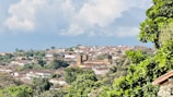 A picturesque landscape of a historic town with lush greenery. The town features traditional colonial architecture, including a prominent church with a dome and bell tower. Whitewashed buildings with red-tiled roofs are nestled among abundant trees and vegetation. The sky is partly cloudy, adding a serene backdrop to the scene.