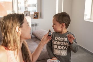 A woman holding a small bottle is interacting with a young child wearing a shirt that reads 'Dad's Gaming Buddy' with pixelated designs. They are indoors with a couch and some framed photos visible in the background.