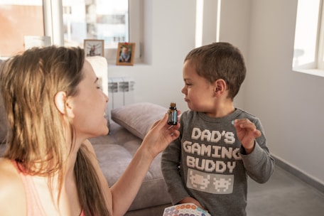 A woman holding a small bottle is interacting with a young child wearing a shirt that reads 'Dad's Gaming Buddy' with pixelated designs. They are indoors with a couch and some framed photos visible in the background.