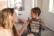 A woman holding a small bottle is interacting with a young child wearing a shirt that reads 'Dad's Gaming Buddy' with pixelated designs. They are indoors with a couch and some framed photos visible in the background.