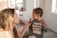 A woman holding a small bottle is interacting with a young child wearing a shirt that reads 'Dad's Gaming Buddy' with pixelated designs. They are indoors with a couch and some framed photos visible in the background.