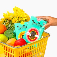 A yellow shopping basket is filled with a variety of fruits and vegetables, including oranges, apples, and broccoli. Bright yellow flowers are also present. Two turquoise boxes of Joolies organic Medjool dates are prominently displayed, one being held by a hand with painted nails.
