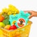 A yellow shopping basket is filled with a variety of fruits and vegetables, including oranges, apples, and broccoli. Bright yellow flowers are also present. Two turquoise boxes of Joolies organic Medjool dates are prominently displayed, one being held by a hand with painted nails.