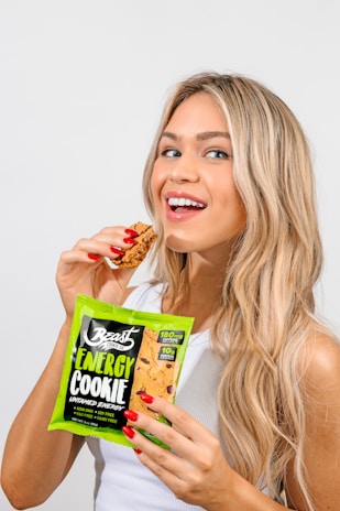 A woman with long blonde hair is smiling and holding a packaged energy cookie in one hand while taking a bite from a cookie piece in the other hand. She's wearing a white sleeveless top and has red nail polish.