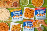An assortment of snacks is arranged on a green tablecloth with a football field pattern. Displayed are bags of Snow Days Pizza Bites in three flavors, surrounded by bowls of popcorn, pretzels, potato chips, nuts, a slice of pizza, and a bowl of guacamole. A person's hands are visible holding a small plate with pizza bites, tortilla chips, and a dipping sauce.