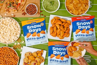 An assortment of snacks is arranged on a green tablecloth with a football field pattern. Displayed are bags of Snow Days Pizza Bites in three flavors, surrounded by bowls of popcorn, pretzels, potato chips, nuts, a slice of pizza, and a bowl of guacamole. A person's hands are visible holding a small plate with pizza bites, tortilla chips, and a dipping sauce.