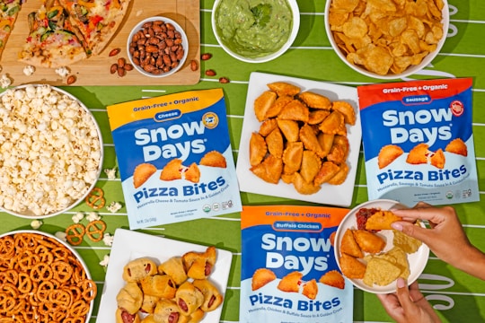 An assortment of snacks is arranged on a green tablecloth with a football field pattern. Displayed are bags of Snow Days Pizza Bites in three flavors, surrounded by bowls of popcorn, pretzels, potato chips, nuts, a slice of pizza, and a bowl of guacamole. A person's hands are visible holding a small plate with pizza bites, tortilla chips, and a dipping sauce.