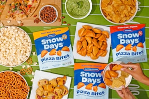 An assortment of snacks is arranged on a green tablecloth with a football field pattern. Displayed are bags of Snow Days Pizza Bites in three flavors, surrounded by bowls of popcorn, pretzels, potato chips, nuts, a slice of pizza, and a bowl of guacamole. A person's hands are visible holding a small plate with pizza bites, tortilla chips, and a dipping sauce.