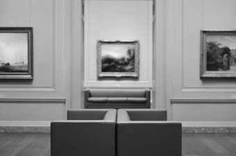 A monochrome image of an art gallery with framed paintings on the walls. There is a leather bench in the foreground and another in the background between two doorways. The paintings appear to depict landscapes and are hung in ornate frames on paneled walls. The room has a classic and elegant design.