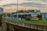 City buses travel on a raised highway with a guardrail bordered by a multi-colored pattern, under a cloudy sky at dusk. In the background, urban buildings with varied architecture are visible, including a hotel and possible mosque with minarets. Streetlights line the road, casting subtle glows as motorbikes cruise below.