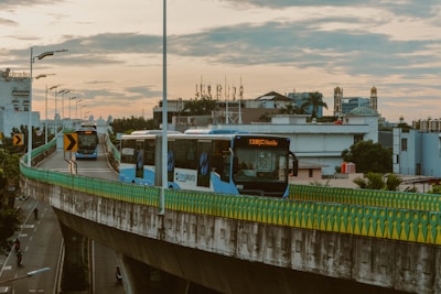 City buses travel on a raised highway with a guardrail bordered by a multi-colored pattern, under a cloudy sky at dusk. In the background, urban buildings with varied architecture are visible, including a hotel and possible mosque with minarets. Streetlights line the road, casting subtle glows as motorbikes cruise below.