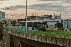City buses travel on a raised highway with a guardrail bordered by a multi-colored pattern, under a cloudy sky at dusk. In the background, urban buildings with varied architecture are visible, including a hotel and possible mosque with minarets. Streetlights line the road, casting subtle glows as motorbikes cruise below.