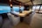 A luxurious interior space featuring a large, elegant bar counter surrounded by plush, comfortable armchairs. The room is well-lit with soft lighting fixtures above, and large windows provide a clear view of a scenic ocean vista. The decor feels sophisticated and upscale with polished wood and neutral tones. A bouquet of flowers can be seen on the far end of the counter, adding a touch of freshness to the setting.