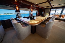 A luxurious interior space featuring a large, elegant bar counter surrounded by plush, comfortable armchairs. The room is well-lit with soft lighting fixtures above, and large windows provide a clear view of a scenic ocean vista. The decor feels sophisticated and upscale with polished wood and neutral tones. A bouquet of flowers can be seen on the far end of the counter, adding a touch of freshness to the setting.