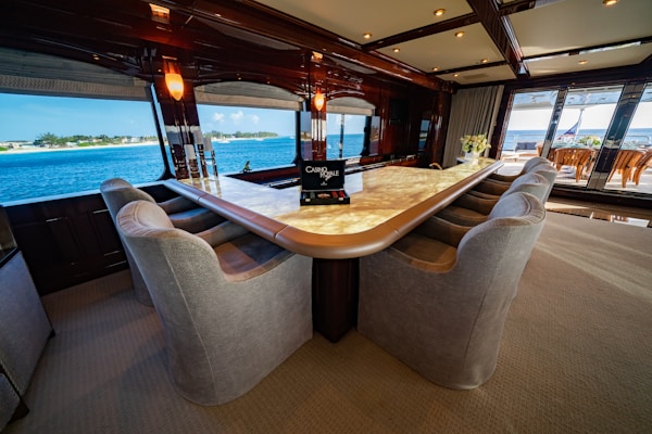 A luxurious interior space featuring a large, elegant bar counter surrounded by plush, comfortable armchairs. The room is well-lit with soft lighting fixtures above, and large windows provide a clear view of a scenic ocean vista. The decor feels sophisticated and upscale with polished wood and neutral tones. A bouquet of flowers can be seen on the far end of the counter, adding a touch of freshness to the setting.
