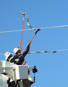 Two utility workers are in a bucket lift, wearing protective helmets and gloves, using long insulated poles to work on power lines against a clear blue sky.