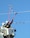 Two utility workers are in a bucket lift, wearing protective helmets and gloves, using long insulated poles to work on power lines against a clear blue sky.