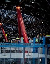 The image depicts a spacious indoor area with red structural pillars reaching up to a complex ceiling grid of metal beams and lights. The reflective flooring is lined with blue barriers, forming guided pathways. In the background, various signs and kiosks are visible, suggesting a busy, public setting like an airport or a convention center.