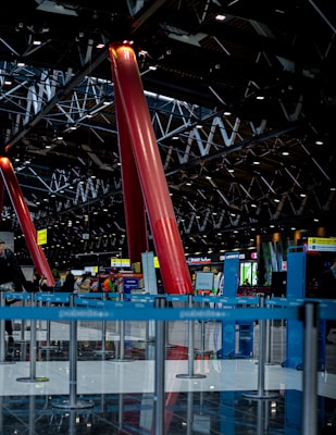 The image depicts a spacious indoor area with red structural pillars reaching up to a complex ceiling grid of metal beams and lights. The reflective flooring is lined with blue barriers, forming guided pathways. In the background, various signs and kiosks are visible, suggesting a busy, public setting like an airport or a convention center.