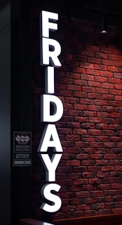 Large, illuminated white letters spelling 'FRIDAYS' are mounted vertically on a textured red brick wall. Below is a sign with information written in both English and another language, possibly indicating a bar or restaurant with various logos and text.