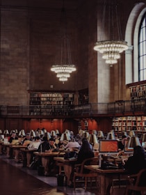 A grand library reading room filled with people working on computers and reading at long wooden tables. The room features high ceilings, large chandeliers, and an expansive arched window. Bookshelves line the walls, filled with numerous books.