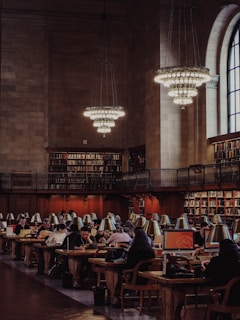 A grand library reading room filled with people working on computers and reading at long wooden tables. The room features high ceilings, large chandeliers, and an expansive arched window. Bookshelves line the walls, filled with numerous books.