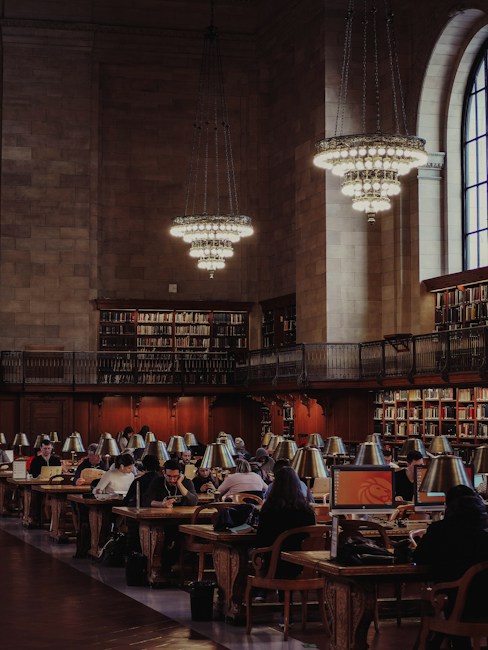 A grand library reading room filled with people working on computers and reading at long wooden tables. The room features high ceilings, large chandeliers, and an expansive arched window. Bookshelves line the walls, filled with numerous books.