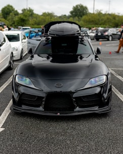 A sleek black sports car parked in a lot, featuring modern headlights and a roof box. Several other vehicles and people are visible in the background, along with overcast skies and trees.
