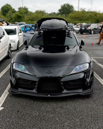 A sleek black sports car parked in a lot, featuring modern headlights and a roof box. Several other vehicles and people are visible in the background, along with overcast skies and trees.