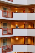 A person is standing on a white balcony with burnt orange railings, holding a piece of paper. The building has multiple floors with similar balconies and warm, yellow lighting illuminating the hallways. The decor suggests a hotel or an apartment building.