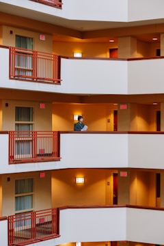 A person is standing on a white balcony with burnt orange railings, holding a piece of paper. The building has multiple floors with similar balconies and warm, yellow lighting illuminating the hallways. The decor suggests a hotel or an apartment building.
