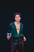 A person stands confidently in leather pants and an unbuttoned green shirt, revealing a prominent tattoo of a butterfly on the chest. The individual is holding a microphone with a logo, and the background is dark, suggesting a stage or event setting.