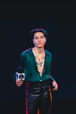 A person stands confidently in leather pants and an unbuttoned green shirt, revealing a prominent tattoo of a butterfly on the chest. The individual is holding a microphone with a logo, and the background is dark, suggesting a stage or event setting.