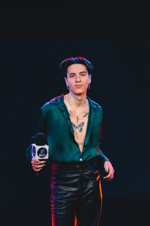 A person stands confidently in leather pants and an unbuttoned green shirt, revealing a prominent tattoo of a butterfly on the chest. The individual is holding a microphone with a logo, and the background is dark, suggesting a stage or event setting.